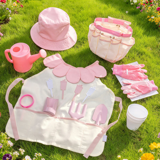Light pink children's gardening set flat-lay on grass: tote bag, sun hat, apron, gloves, watering can and tools