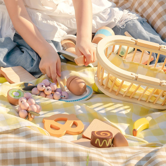 ROBUD Kids Wooden Picnic Play Food Set outdoor lifestyle with child hands arranging banana, cookie and wooden food on yellow checked picnic blanket