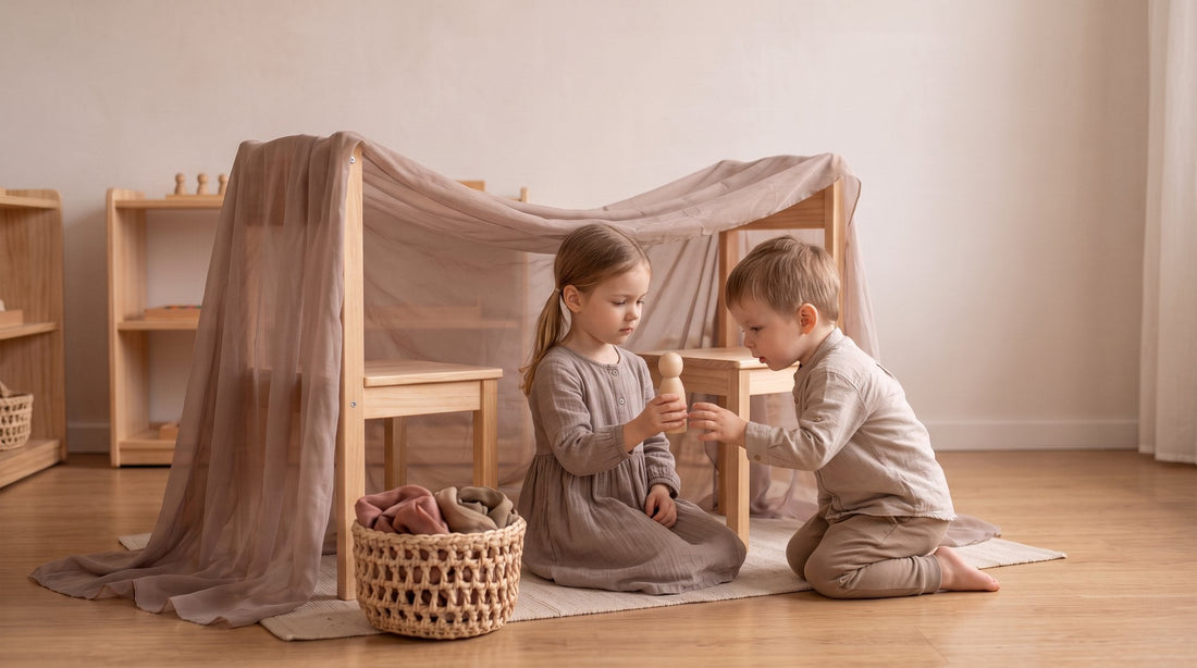 Two children, about four and six years old, playing inside a den made from a sheer play silk draped over two wooden chairs. The older child kneels at the opening holding up a wooden peg doll to show the younger child sitting inside on a cushion.