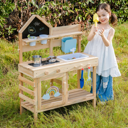 Girl reaching for a yellow toy shovel beside the outdoor wooden mud kitchen, garden setting