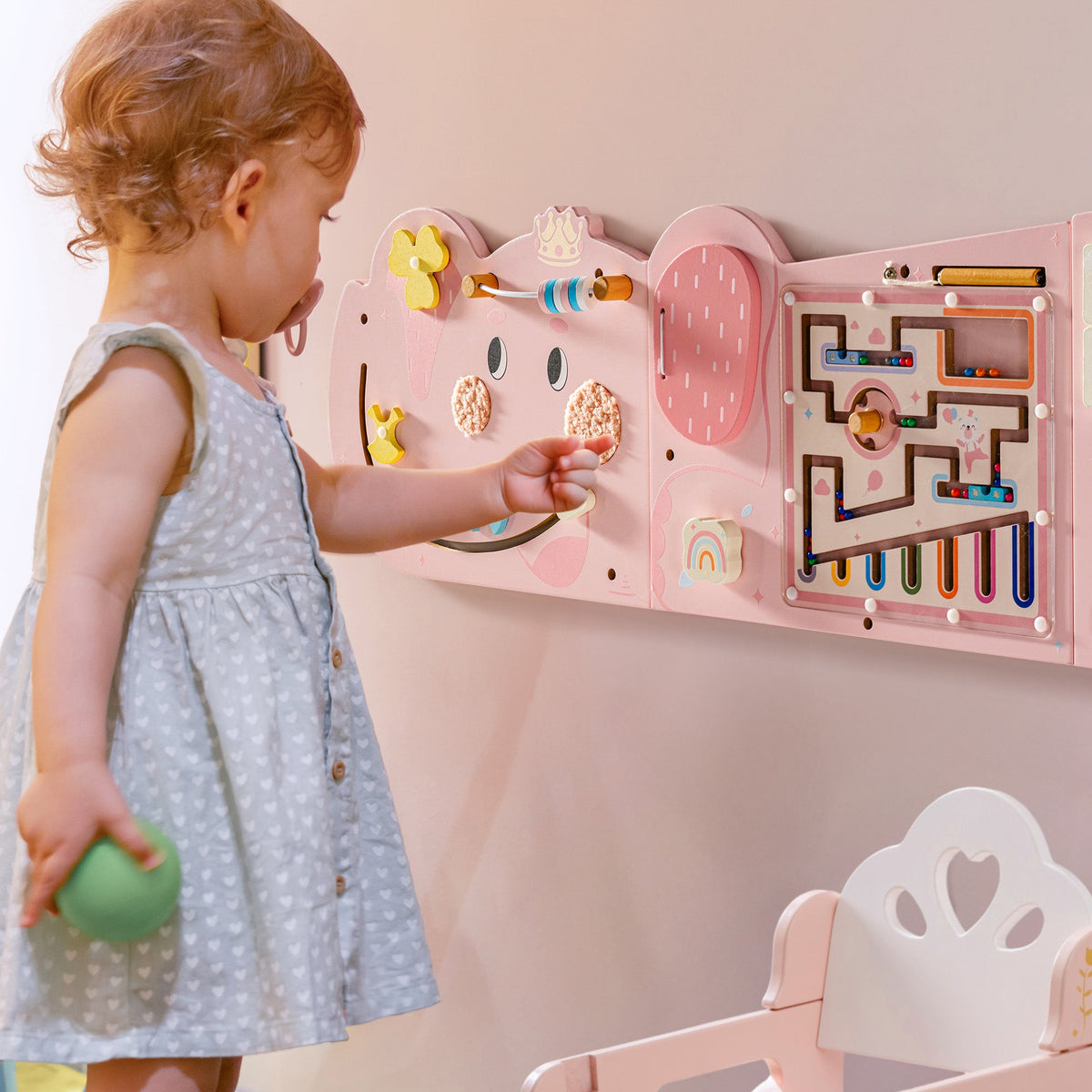 Toddler girl reaching to touch the pink elephant activity busy board mounted on the wall