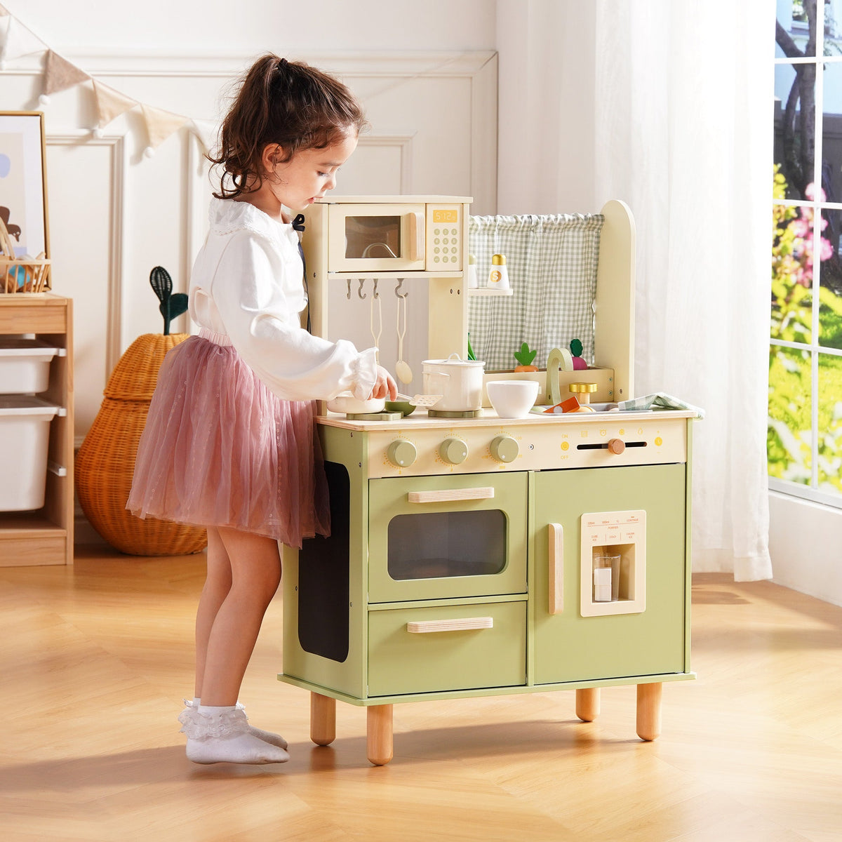 Girl in a pink tutu stands at the sage green wooden play kitchen and opens the refrigerator door compartment.