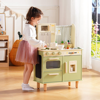 Girl in a pink tutu stands at the sage green wooden play kitchen and opens the refrigerator door compartment.