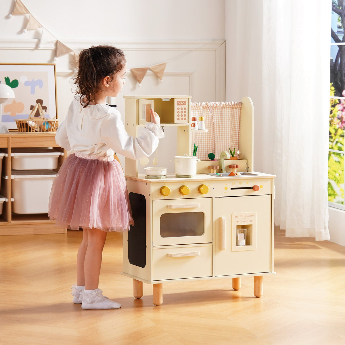 Girl in a pink tutu stands at the cream wooden play kitchen, reaching toward the oven and refrigerator from the side.
