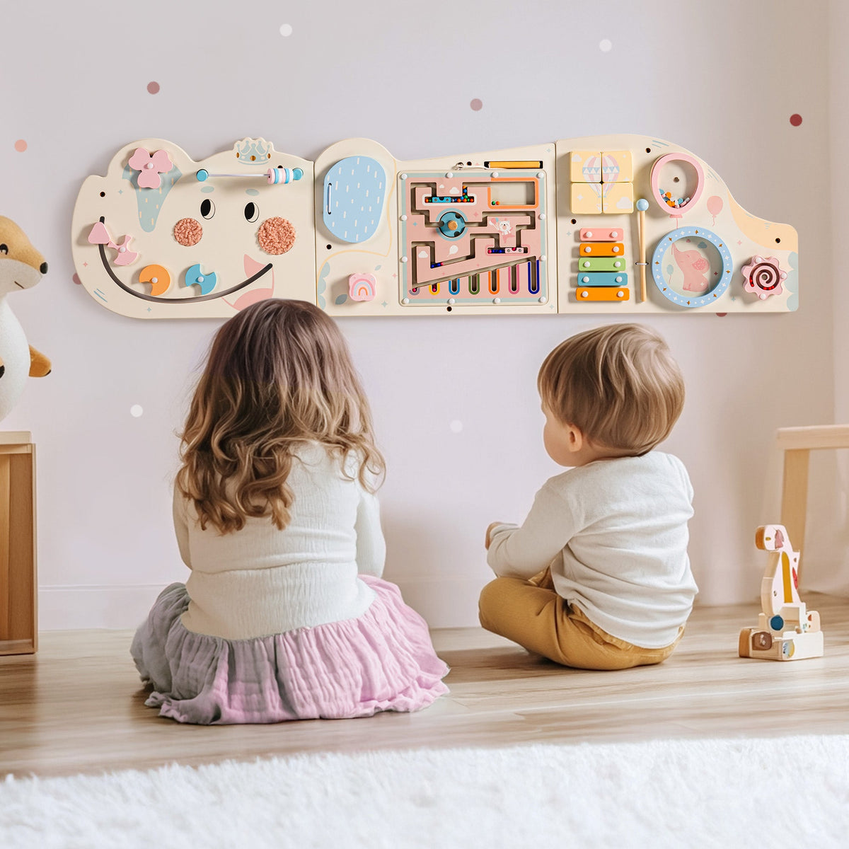 Two children sitting on the floor with their backs to the wall-mounted cream elephant busy board
