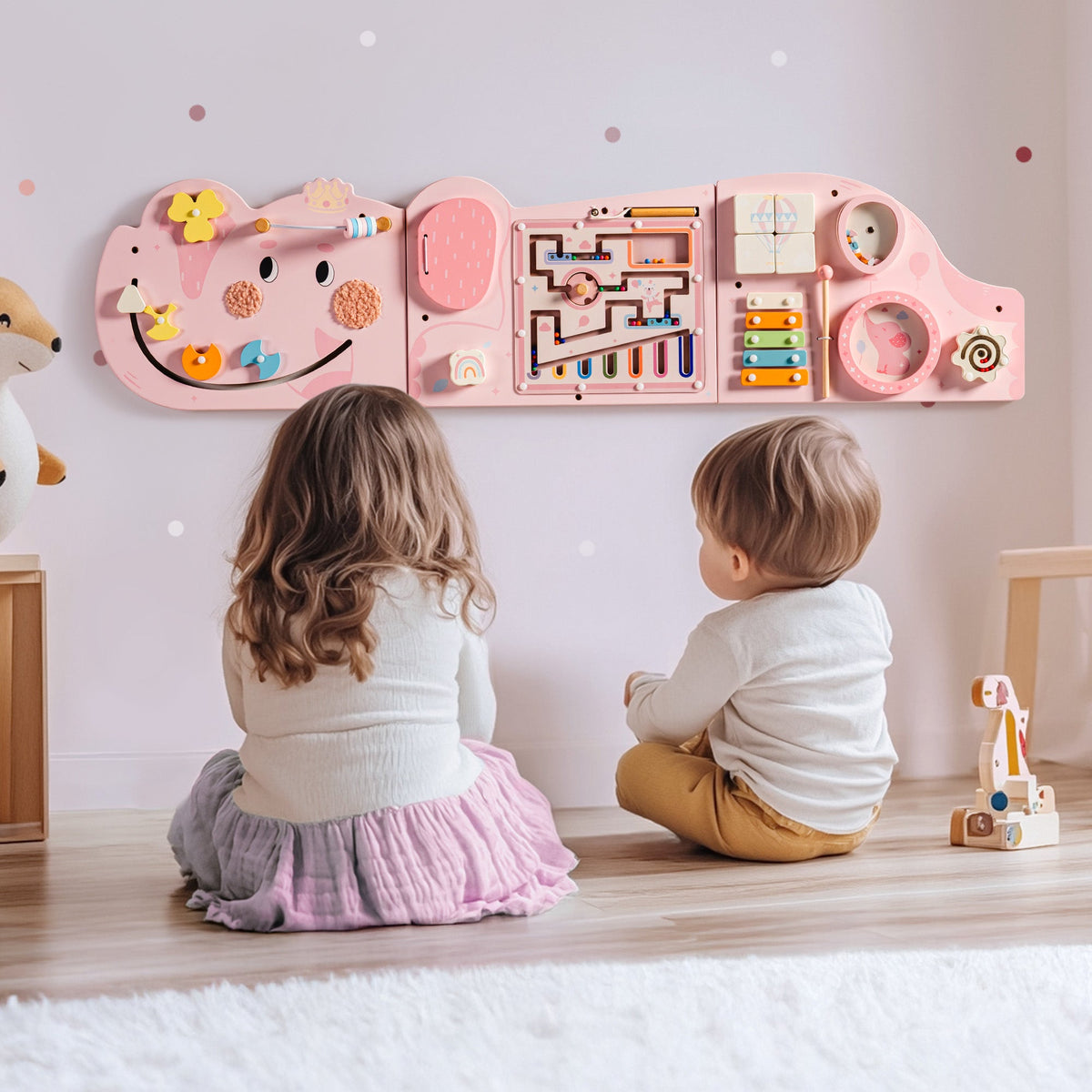 Two children sitting on the floor with their backs to the wall-mounted pink elephant busy board