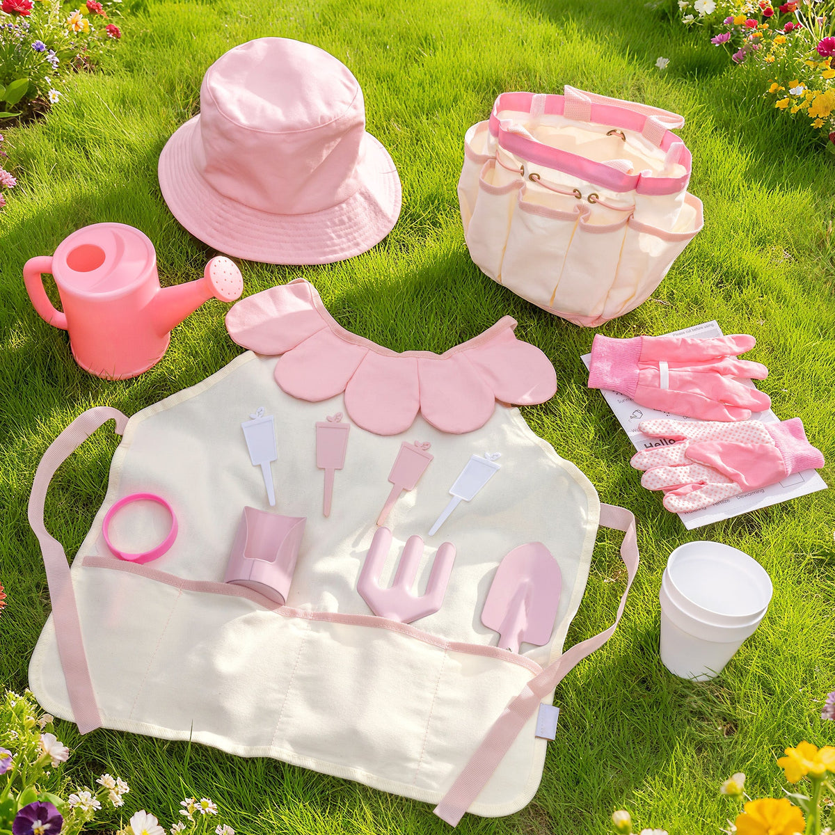 Light pink children's gardening set flat-lay on grass: tote bag, sun hat, apron, gloves, watering can and tools