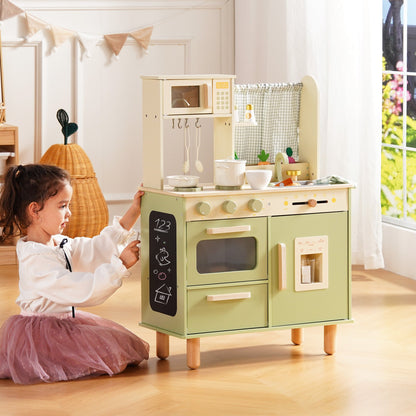 Girl in a tutu kneels beside the sage green wooden play kitchen, writing on the chalkboard panel.