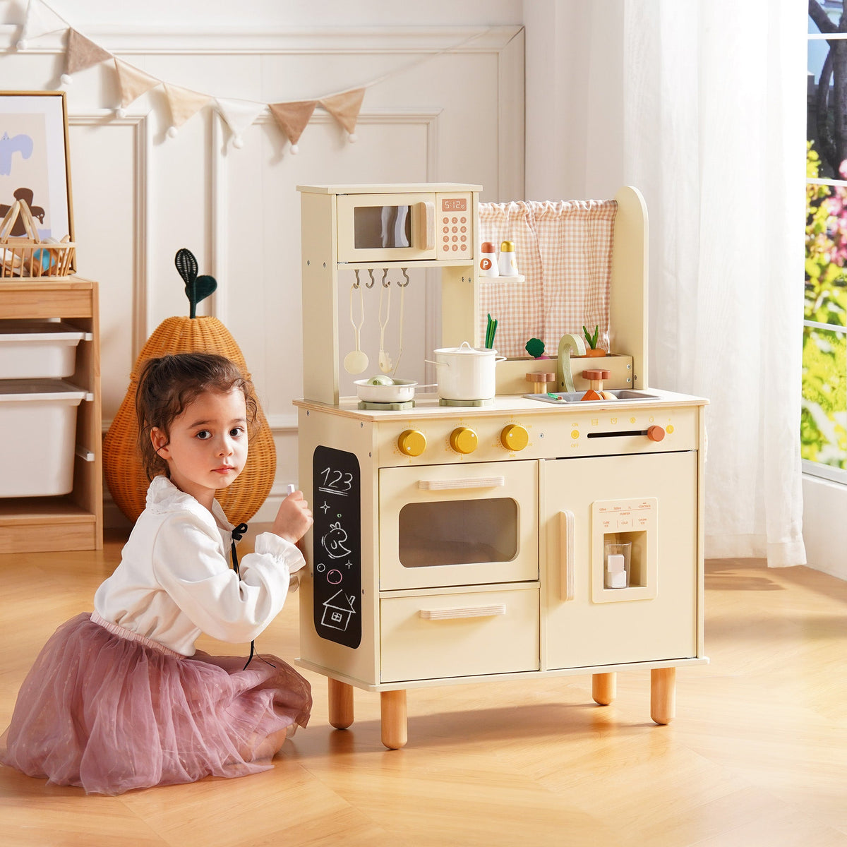 Girl in a tutu kneels at floor level beside the cream wooden play kitchen, scribbling on the lower chalkboard panel.