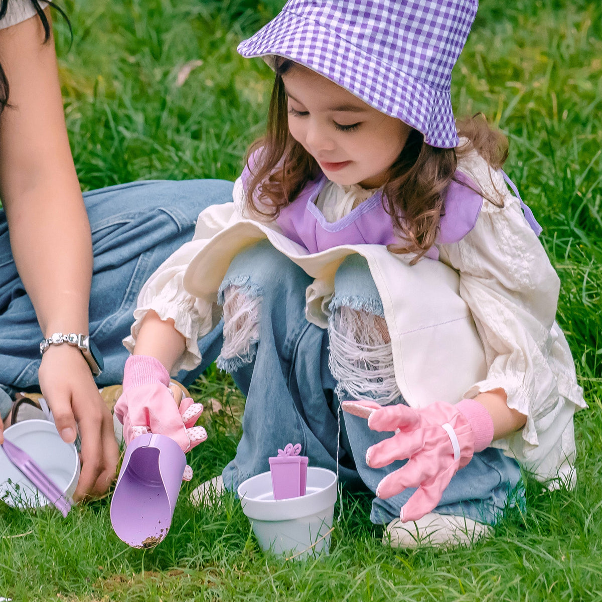 Girl in purple plaid hat peering through the gardening set's magnifying glass outdoors among plants and flowers