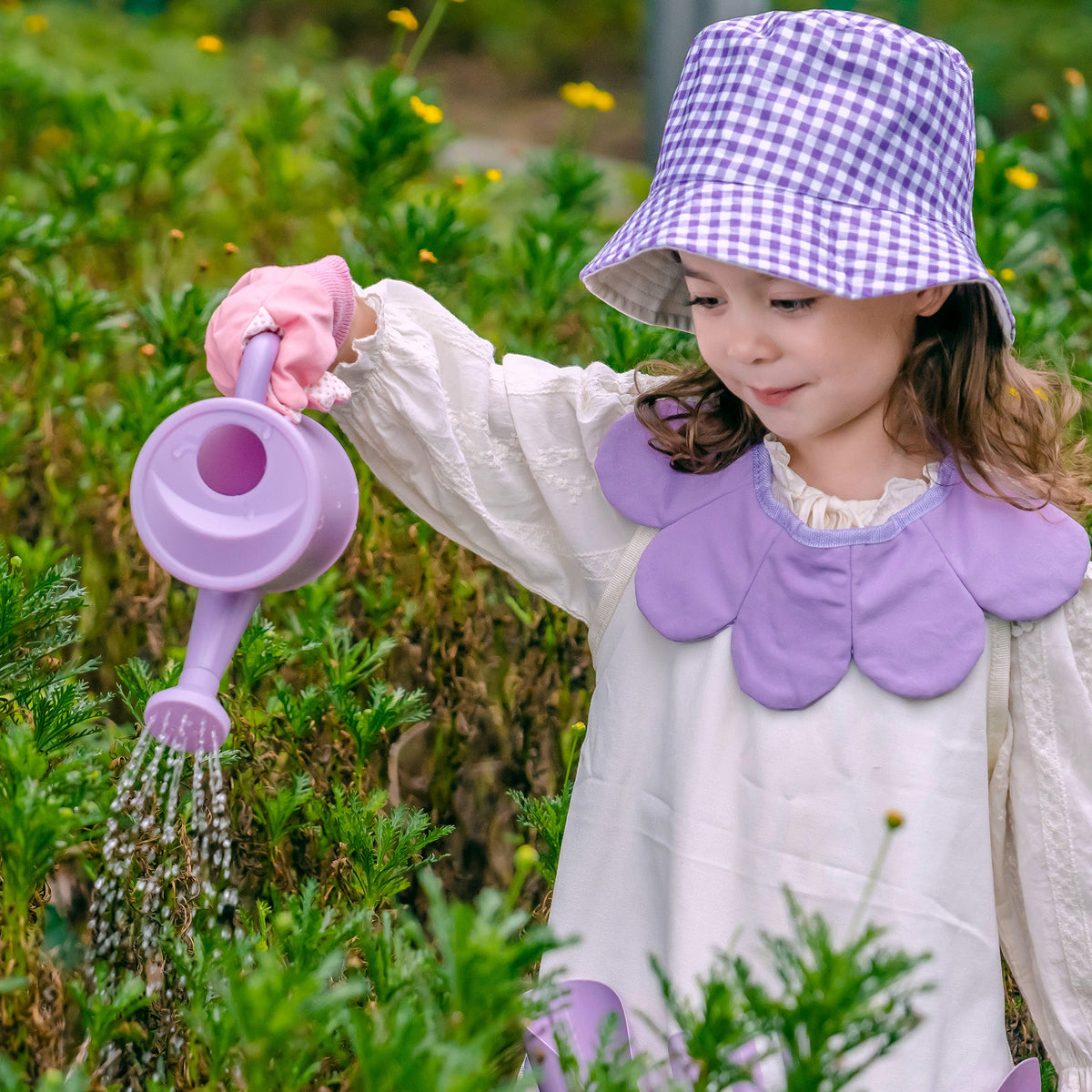 Girl in a purple plaid hat kneeling on grass, pouring water from the purple gardening set's watering can into a bucket