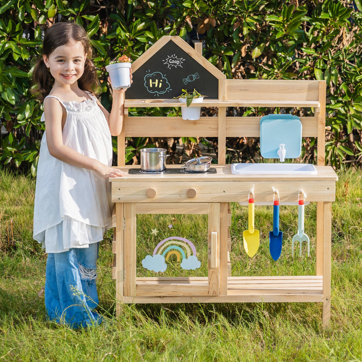 Girl smiling and holding a small terracotta pot beside the outdoor wooden mud kitchen's chalkboard panel, garden setting