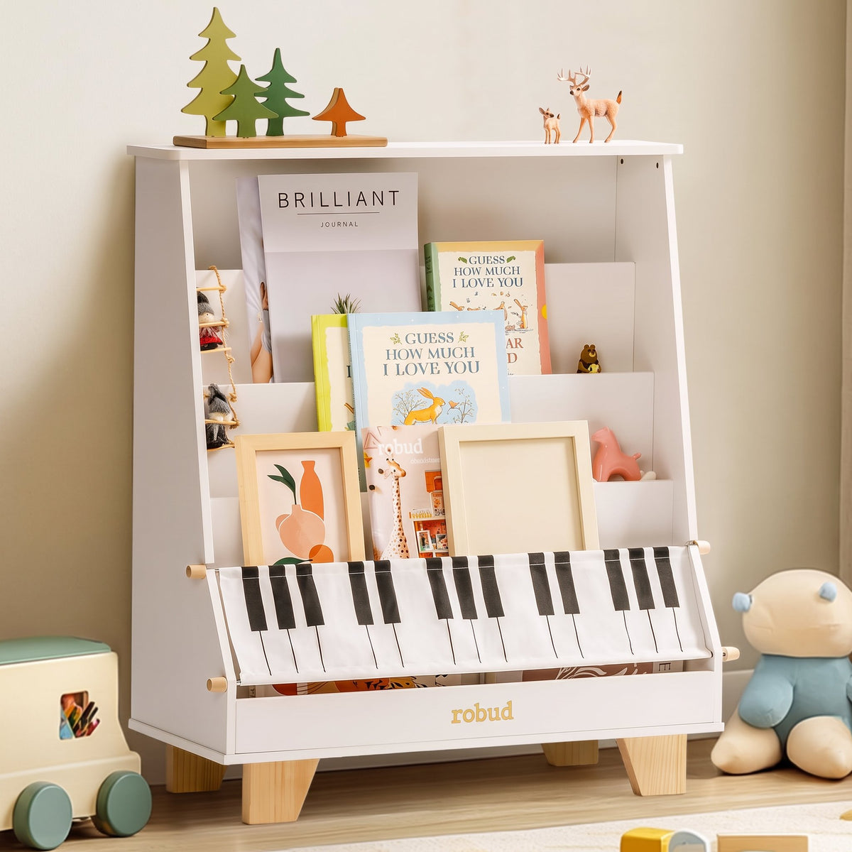 ROBUD wooden kids bookshelf (white) in a room with wooden toys on the floor, showing the tiered front-facing book display and the piano-key fabric storage pocket