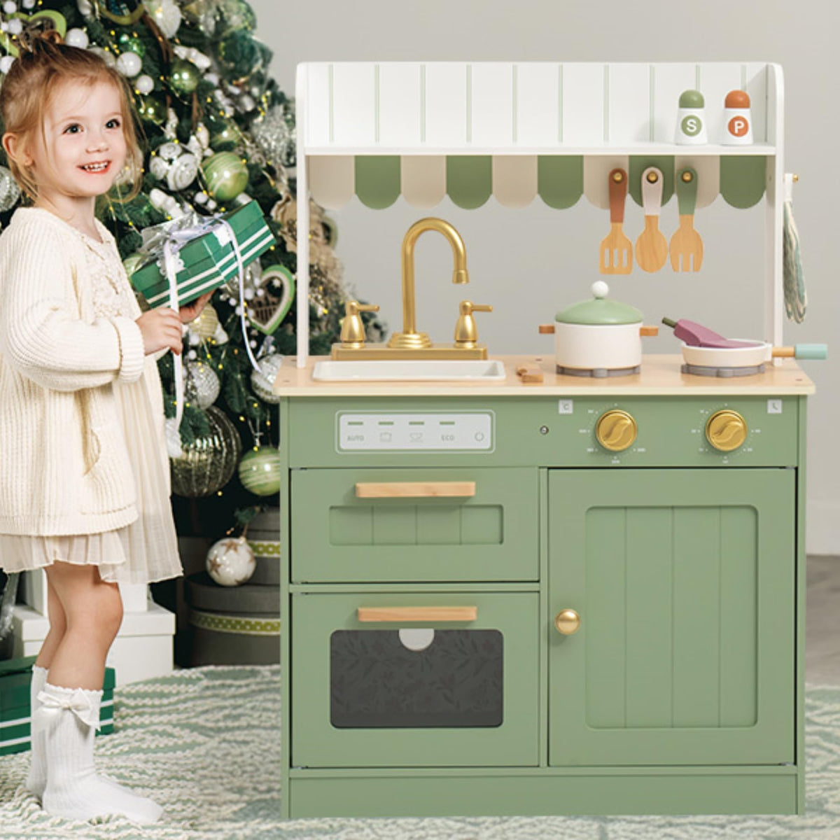 Young girl in a cream dress playing beside the sage green wooden play kitchen set, decorated near a Christmas tree