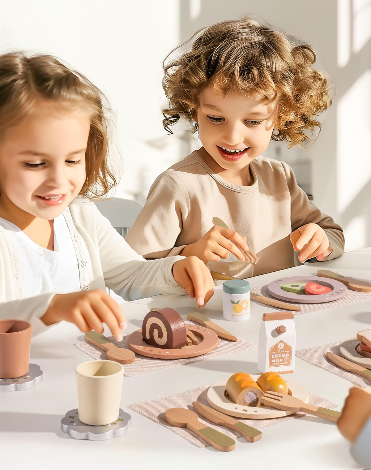 Two children laughing playing tea party with ROBUD wooden play dishes on a sunlit table with cake toy