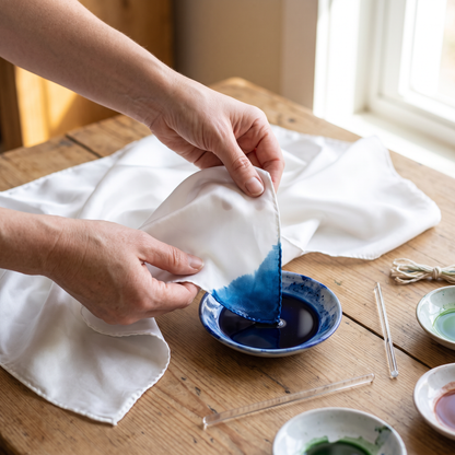Hand dipping the corner of a white silk scarf into blue dye at a home craft table