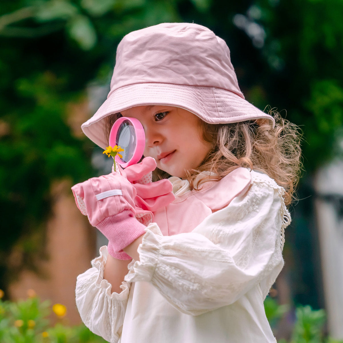 Girl in a light pink sun hat peering through the gardening set's magnifying glass outdoors