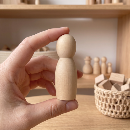 Close-up of an adult's hand holding one natural unfinished wood peg doll, other peg family figures softly out of focus on a shelf behind