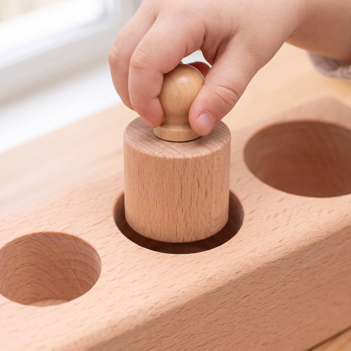 A young child's hand lifting one natural beech knobbed cylinder up out of its socket in a Montessori knobbed cylinder block