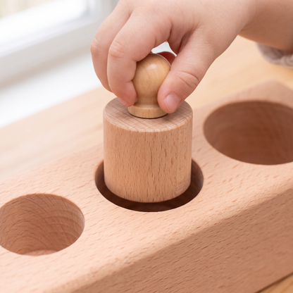 A young child's hand lifting one natural beech knobbed cylinder up out of its socket in a Montessori knobbed cylinder block